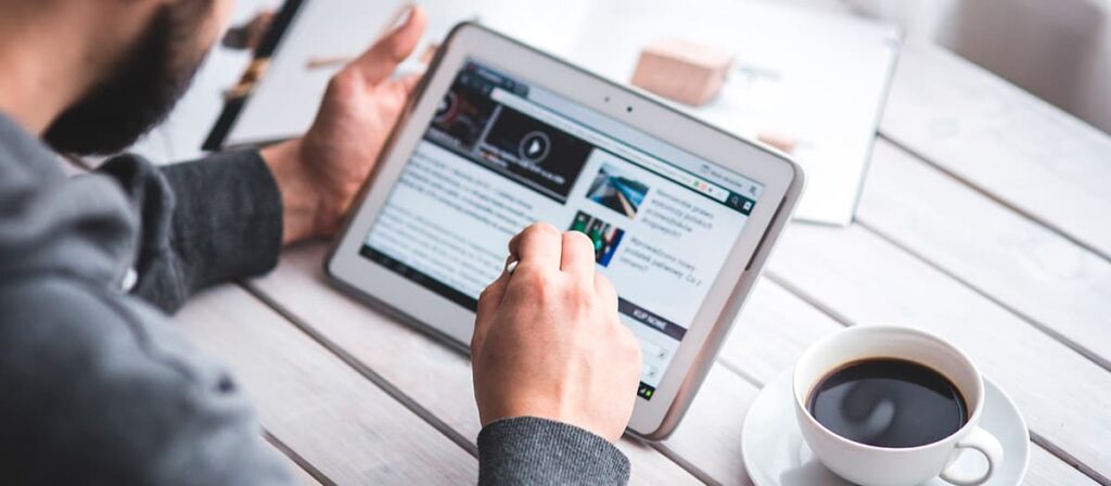 Homem mexendo em um tablet, ao lado dele tem um café.