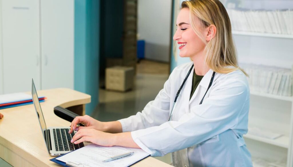Medica em uma sala de consultório, sentada em uma mesa, utilizando um notebook, ela está feliz.