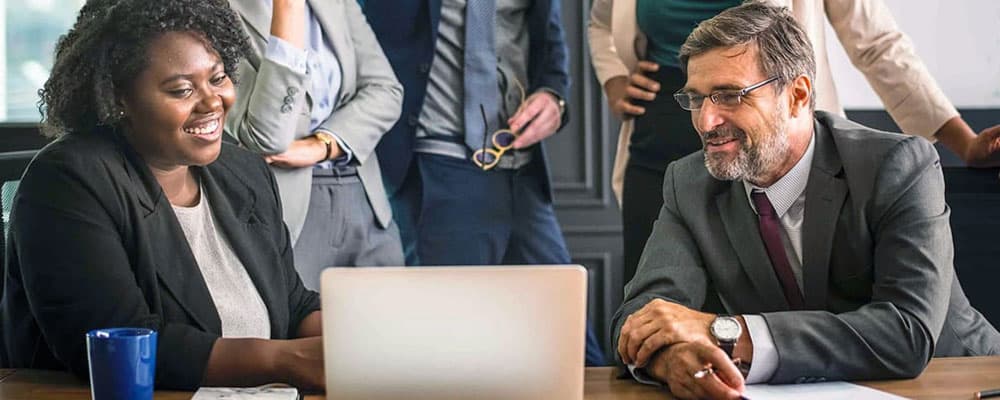 Cinco pessoas reunidas em uma sala de trabalho, estão todos observando algo em um notebook, ambos estão felizes.